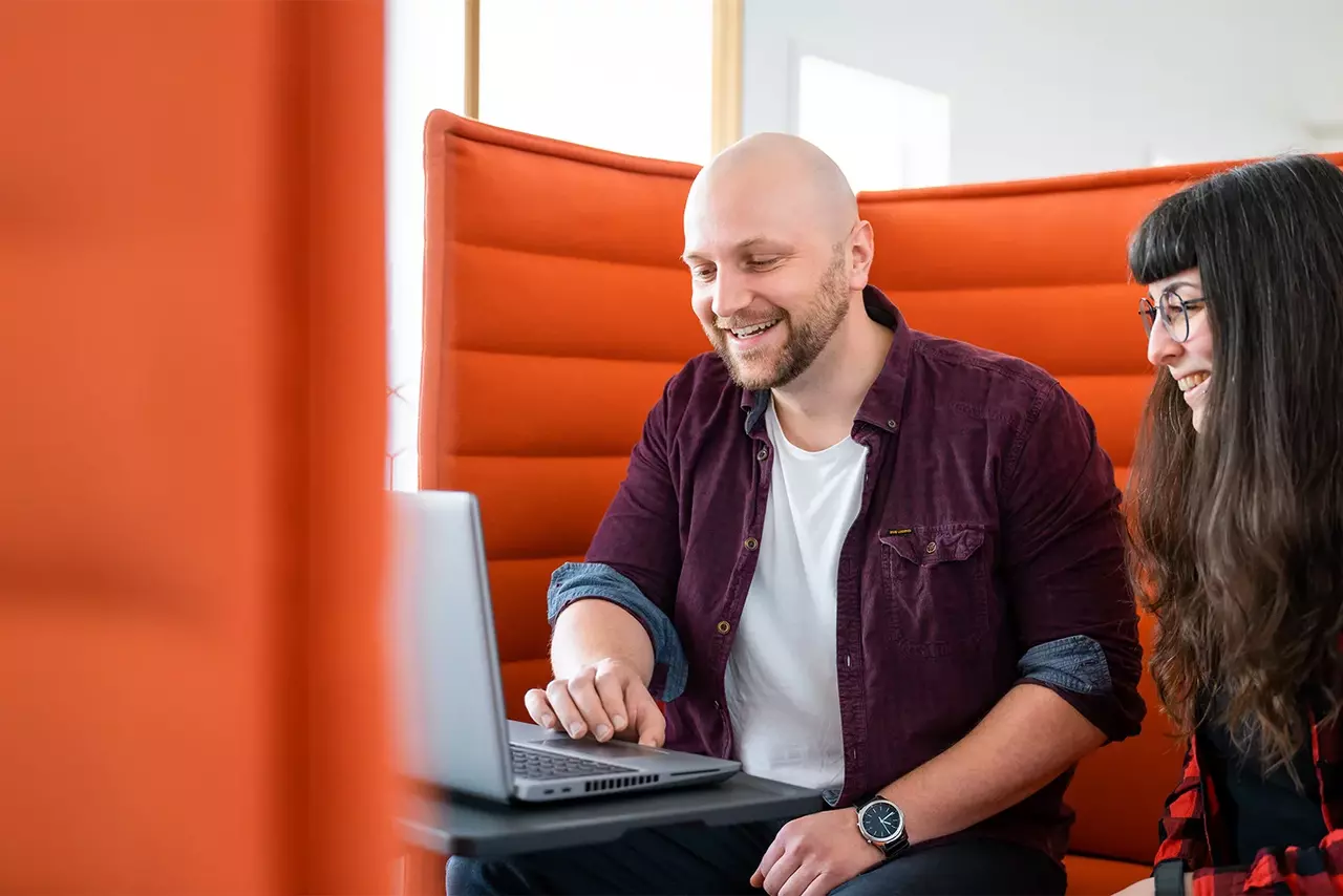 Zwei Mitglieder des Babtec-Teams arbeiten gemeinsam an einem Laptop auf einer orangefarbenen Couch.