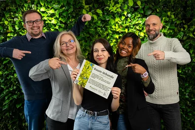 Babtec-Teammitglieder mit der „Baum-Urkunde Zukunftswald“ vor einem blättrig-grünen Hintergrund.